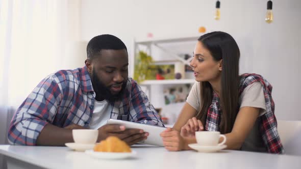 Young Multiethnic Couple Drinking Coffee and Choosing Vacation Trip on Tablet