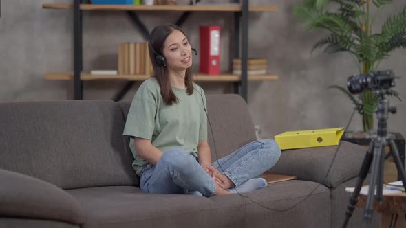 Young Cheerful Asian Woman in Earphones Waving and Talking in Video Camera in Home Office