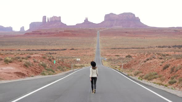 Young woman with hat walking in the middle of famous empty road leading to sandstone buttes of Monum