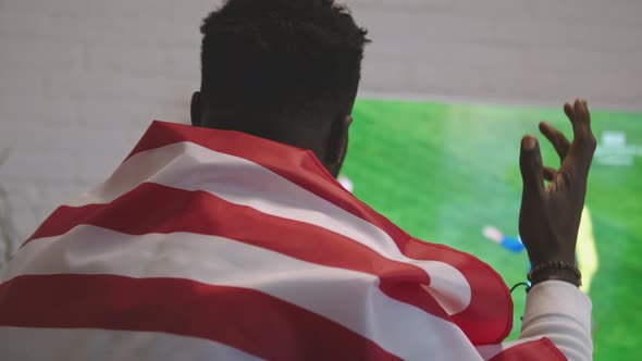 Desperate Black Man with American Flag Over the Shoulders Watching Football Game on Smart Tv