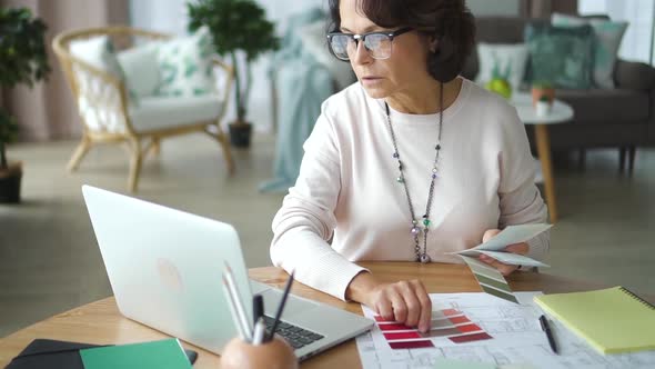 Mature Woman Architect Working with Blueprint at Table with Laptop in Home Room.