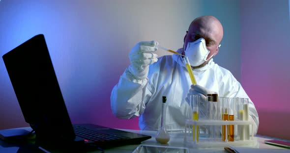 Man in a White Coat, Respirator and Gloves Is Sitting at a Table with a Gadgets. He Holds a Tube