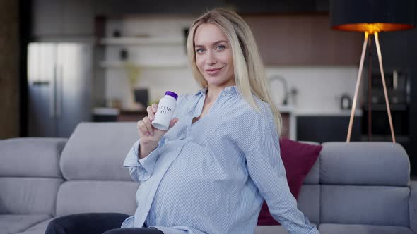 Smiling Confident Pregnant Woman Holding Vitamin D3 Bottle Looking at Camera Sitting on Couch in