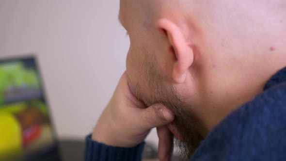 Closeup View From the Back of a Man with a Beard Who Works at a Laptop Watches a Video on the Screen