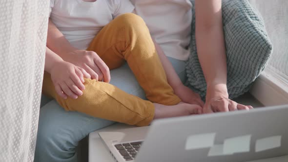 Woman Remote Works From Home. She Sits on Window Sill with Laptop and Cute Toddler Boy on Her Knees