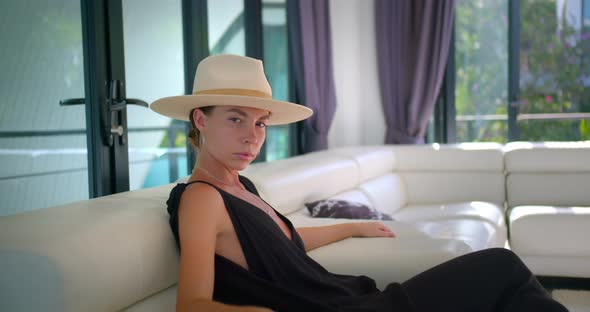 Elegant Woman Seats on the Sofa Indoors She Wears Black Dress and a Hat