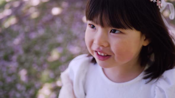 Korean Girl in a White Light Fur Coat and a Headband Stands in a Garden with Cherry Blossoms