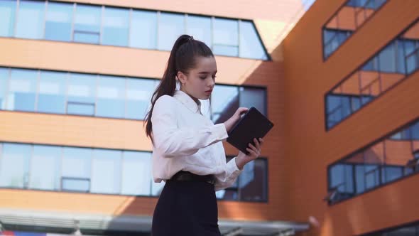 Business Girl Stands Near Business Building Holding a Tablet in Hands