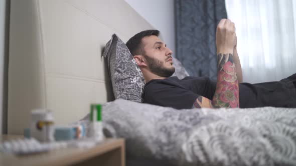 Sick Young Man with Fever Lying in Bed and Checking His Temperature Using Armpit Thermometer