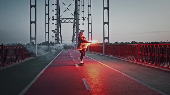 Young Hipster Lady in Informal Outfit is Riding Skateboard on Bridge While Holding Glowing Red