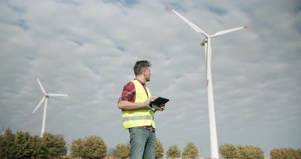 Young Engineer with Tablet in Hand Looks Behind and Works on Wind Mill