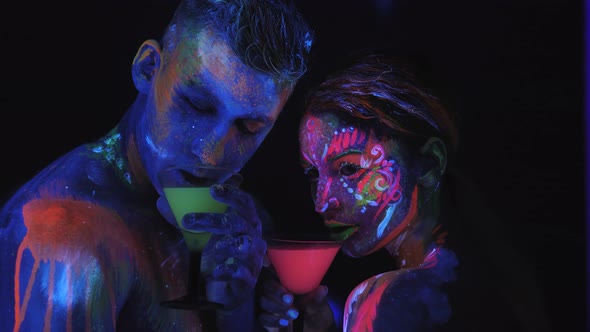 A Couple with UV Body Art with Glasses of Colorful Glowing Cocktails in the Dark