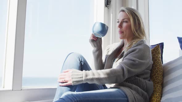 Caucasian mature woman looking through the window and drinking coffee