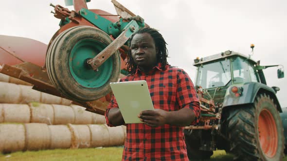 Young African Farmer Using Tablet in Front of Big Green Tractor Anf Haystack