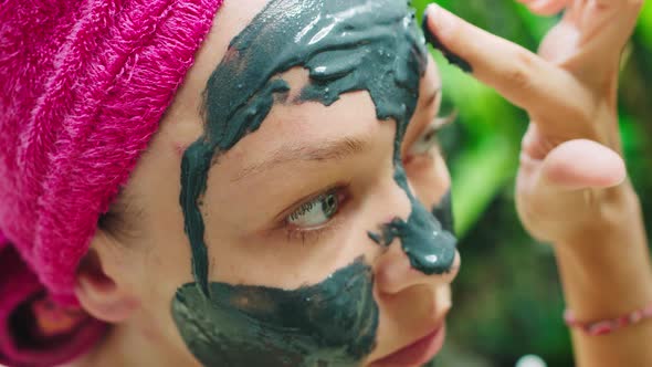 Young woman with towel on head putting face mask on her face after shower in tropical Bali style bat