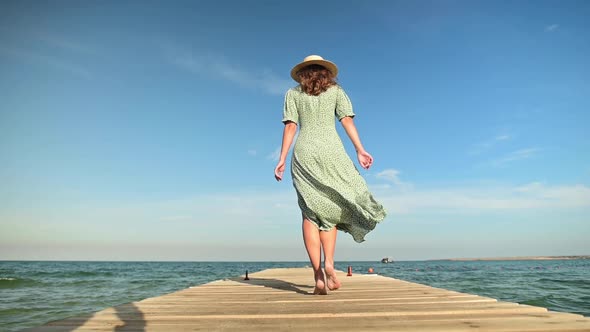 A Lovely Young Caucasian Woman in a Loose Green Dress and a Straw Hat Walks Along a Wooden Pier or