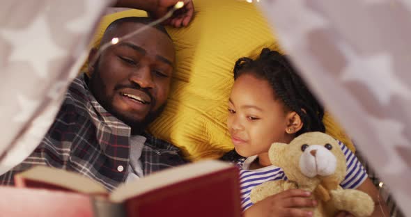 Happy african american father and daughter lying in tent and reading book