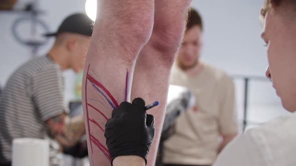 Young Man Tattoo Artist Drawing a Sketch on the Leg on His Client with a Blue Pen