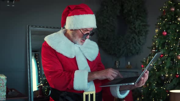 Cheerful Man in Black Glasses in Santa Claus Suit Is Sitting in Living Room and Chatting By Laptop