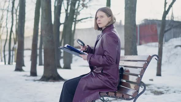 Side View Confident Female Detective Sitting on Bench in Winter Park Examining Evidence with