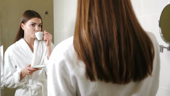 Young Attractive Woman in White Bathrobe Drinking Coffee in Front of Bathroom Mirror