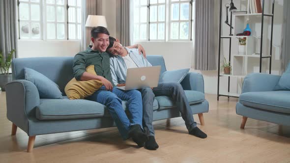 Asian Gentle Gay Couple Using Laptop Computer While Sitting On A Couch In Apartment