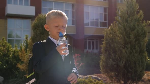 A Small Schoolboy with a Backpack Drinks Water From a Bottle in the Schoolyard