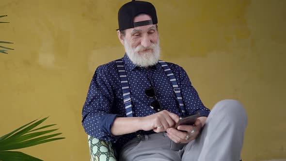 Bearded Senior Man in Casual Clothes and in Stylish Cap Sitting on Chair and Using Phone