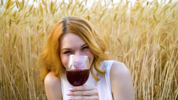 Happy red haired woman drinking wine at picnic in wheat field