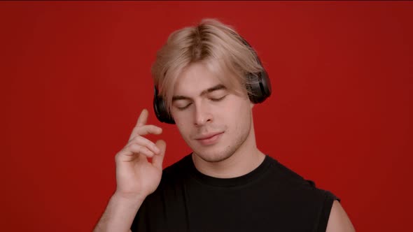 Studio Portrait of Young Blonde Guy Listening To Music in Headphones and Dancing Red Background