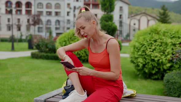 Casual Style Dressed Woman is Resting Outdoors Sitting on Bench in Park and Using Smartphone