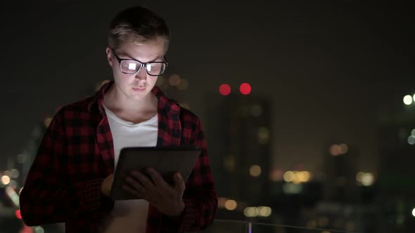 Happy Hipster Man Thinking And Using Digital Tablet Against View Of The City At Night