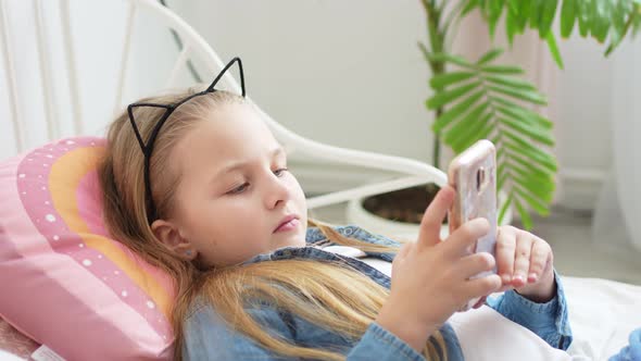 a Beautiful Little Girl in a Rim with Cat Ears with a Phone in Bed