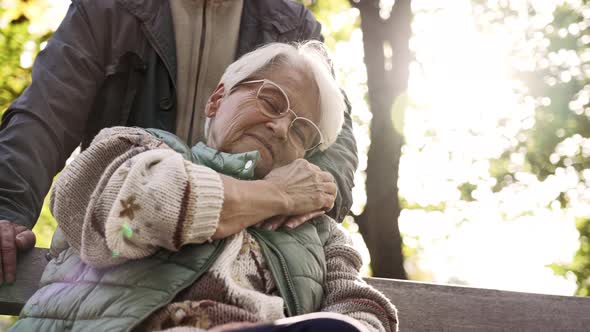 European Caucasian Elderly Lady Sitting on a Bench in a Park Gently Touching Her Husband's Hand