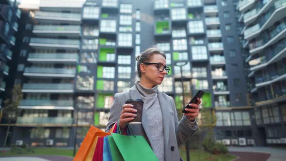 Woman Stands in Business District Holds Takeaway Coffee Shopping Bags and Uses Smartphone