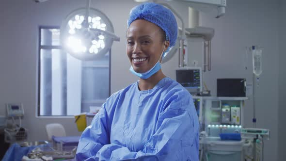 Portrait of mixed race female surgeon wearing lowered face mask smiling in operating theatre