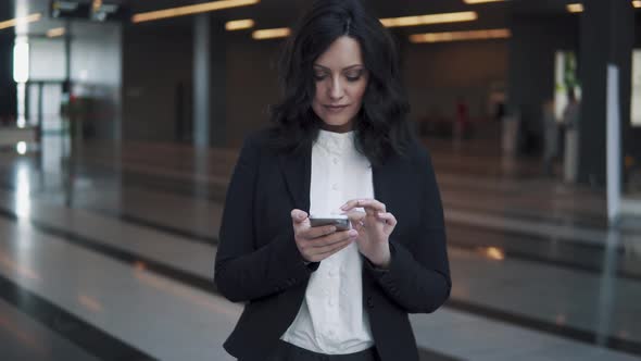 Young Business Woman Uses a Smartphone While Standing in the Lobby of a Modern Office Building