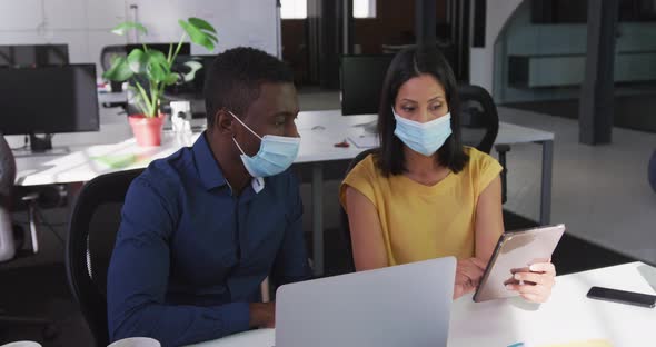 Diverse male and female business colleagues wearing face masks sitting at desk using tablet