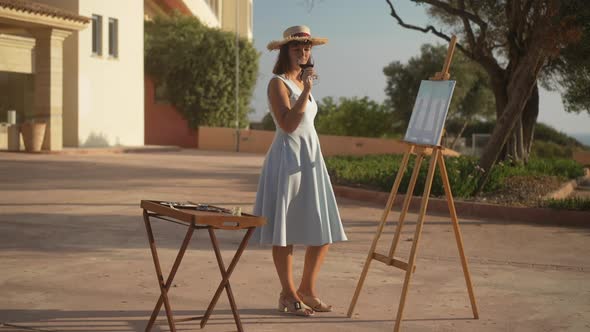 Confident Creative Caucasian Woman in Elegant Dress and Straw Hat Drinking Red Wine Admiring Picture