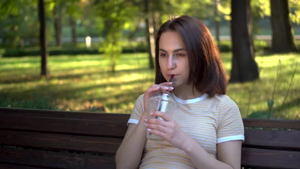 A Young Woman with Glasses Sits on a Bench in the Park and Drinks Cold Coffee Closeup