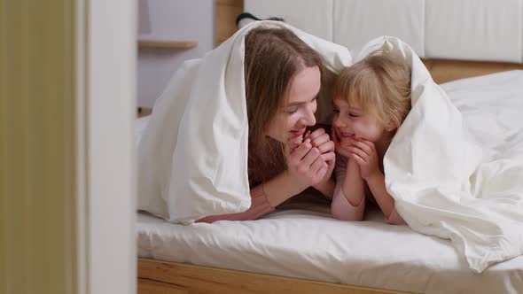 Happy Family of Mother with Daughter Lying on Bed Under Duvet Blanket Telling Secrets to Each Other
