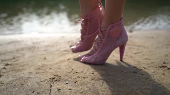 Female Feet Shod Pink Extravaga Fashion Boots, Walking on Sandy Shore of Lake or River
