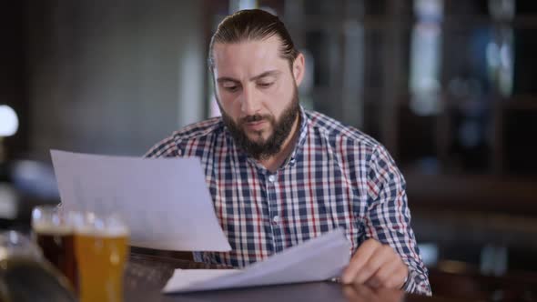 Portrait of Concentrated Caucasian Man Analyzing Paperwork Sitting at Bar Counter Indoors