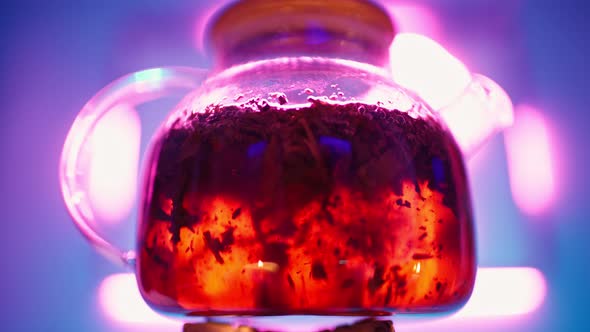 Transparent Teapot with Tea Leaves Boils on Gas Burner Against Neon Backlight