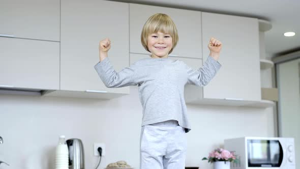 Portrait of Cheerful Caucasian Little Boy Standing in Kitchen Making Strength Gesture Looking at