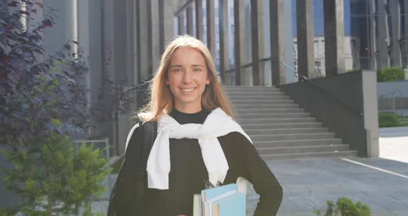 Girl in Casual Clothes with Books Standing in front of City Modern Building and Looking Into Camera