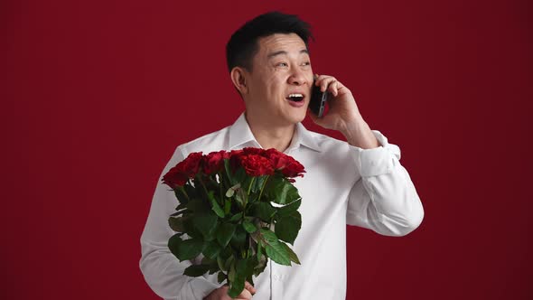 Happy Asian man holding a bouquet of red roses and talking by phone