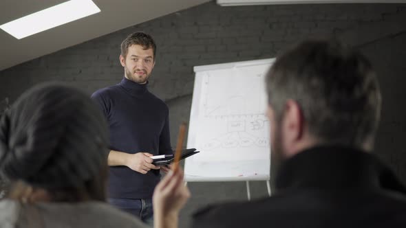 Handsome Young Businessman Pointing at Flipchart During Presentation in Conference Room and Holding