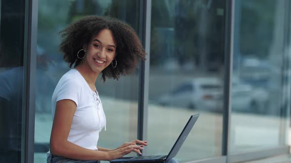 Happy Young Beautiful Afro Woman with Curly Long Hair Enjoys Work on Laptop, Smiling Joyfully in