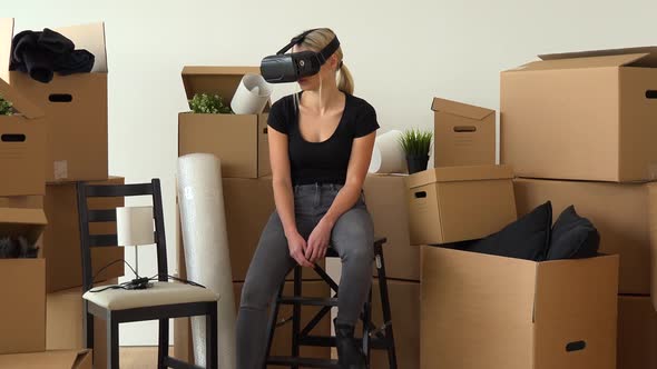 A Moving Woman Sits on a Chair in an Empty Apartment and Uses VR Headset, Surrounded By Boxes